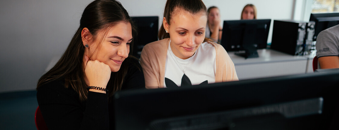 Zwei Schülerinnen sitzen vor dem Computer in der Schule und sehen auf diesen.