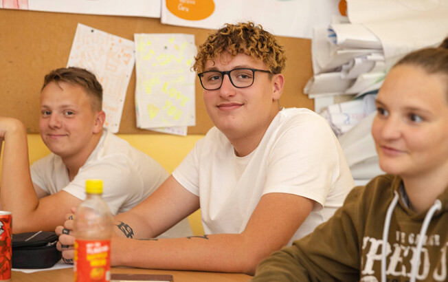Zwei Schüler sitzen im Klassenzimmer und lächeln in die Kamera. Eine Schülerin sitzt noch daneben und sieht zur Tafel.