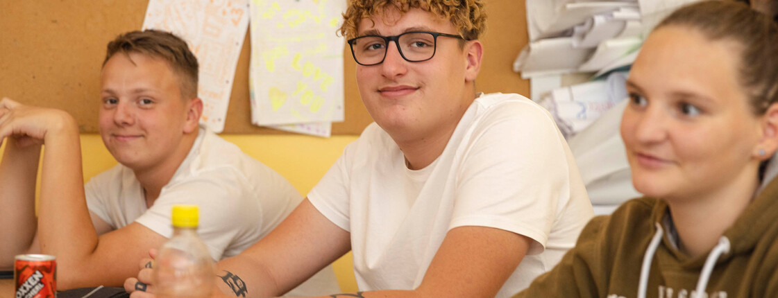 Zwei Schüler sitzen im Klassenzimmer und lächeln in die Kamera. Eine Schülerin sitzt noch daneben und sieht zur Tafel.