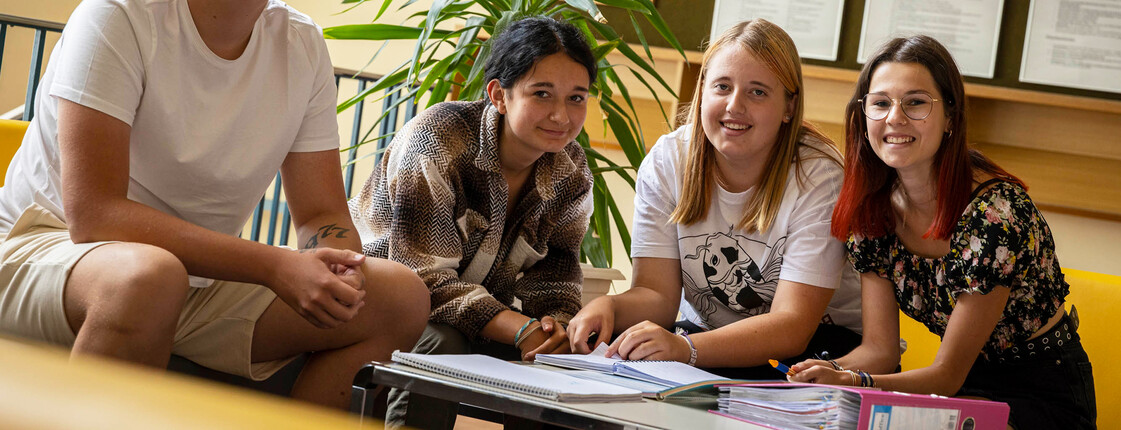 Vier Schüler*innen sitzen auf einem gelben Sofa, haben Schulmaterial vor ihnen liegen und lächeln in die Kamera.