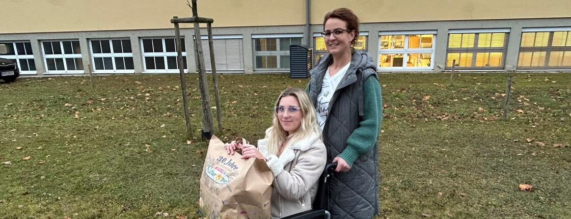 Eine Frau im Rollstuhl hält eine Einkaufstüte am Schoss, während eine weitere Frau den Rollstuhl über den Asphalt schiebt.