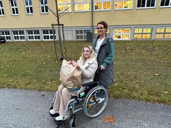 Eine Frau im Rollstuhl hält eine Einkaufstüte am Schoss, während eine weitere Frau den Rollstuhl über den Asphalt schiebt.