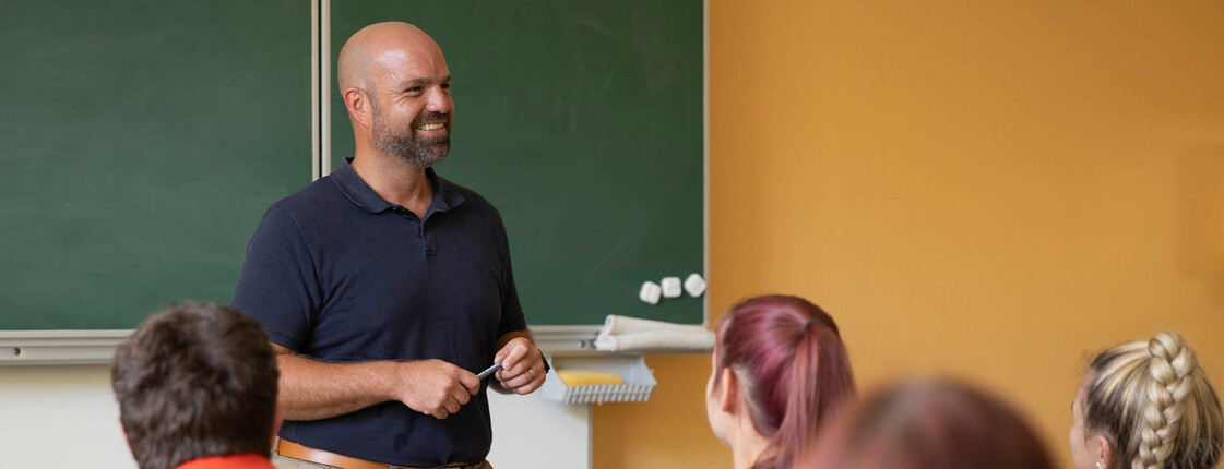 Ein Lehrer steht vorne an der Tafel, die Schüler*innen sehen aufmerksam zu ihm.