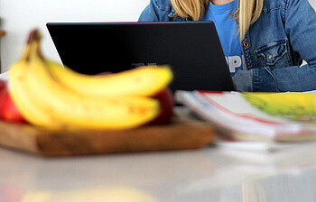 Eine junge Schülerin sitzt vor einem schwarzen Laptop und sieht auf diesen, eine Obstschale und Lernunterlagen liegen vor ihr.
